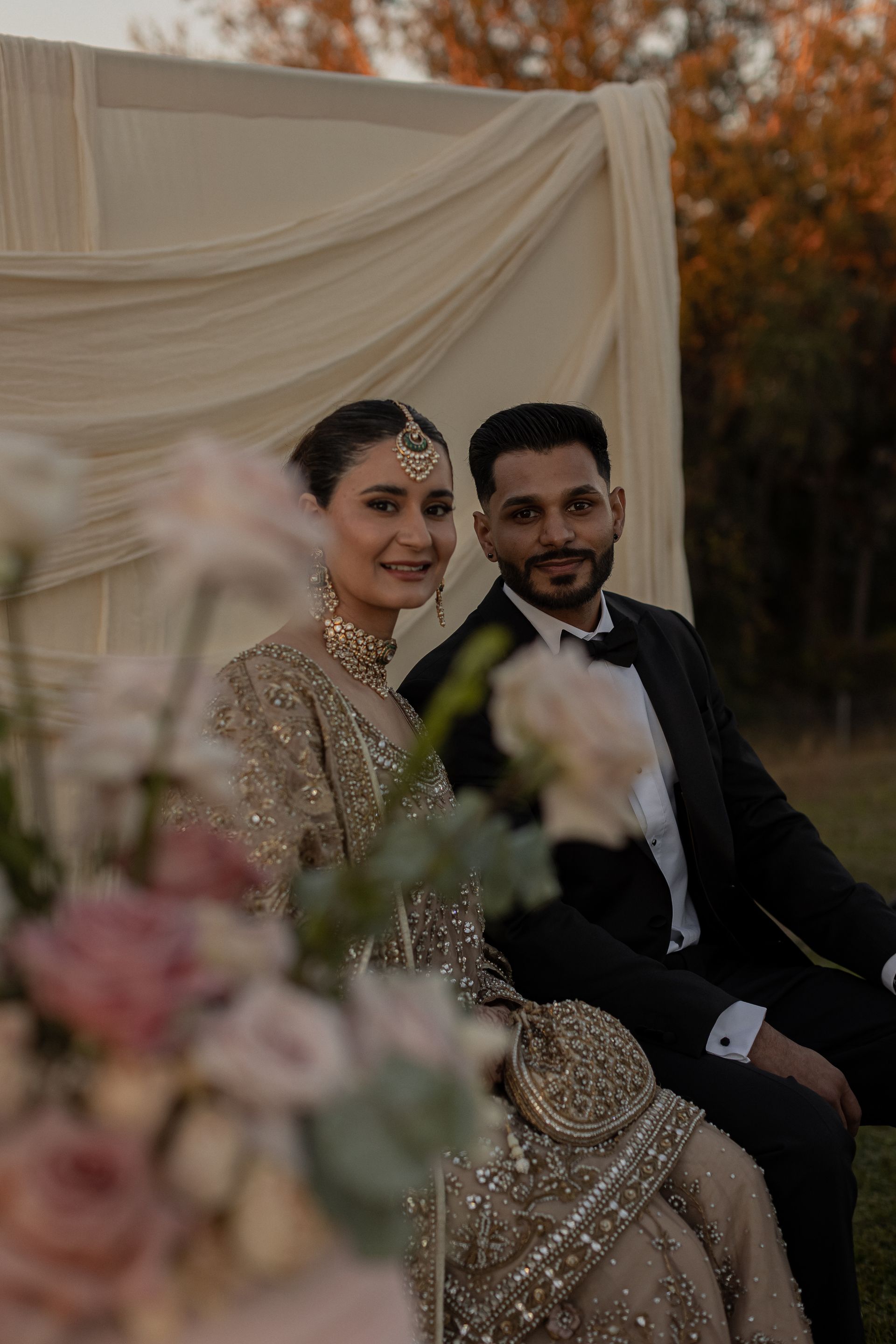 Couple in formal wear, bride in gold, groom in black tuxedo, posing outdoors by draped fabric.