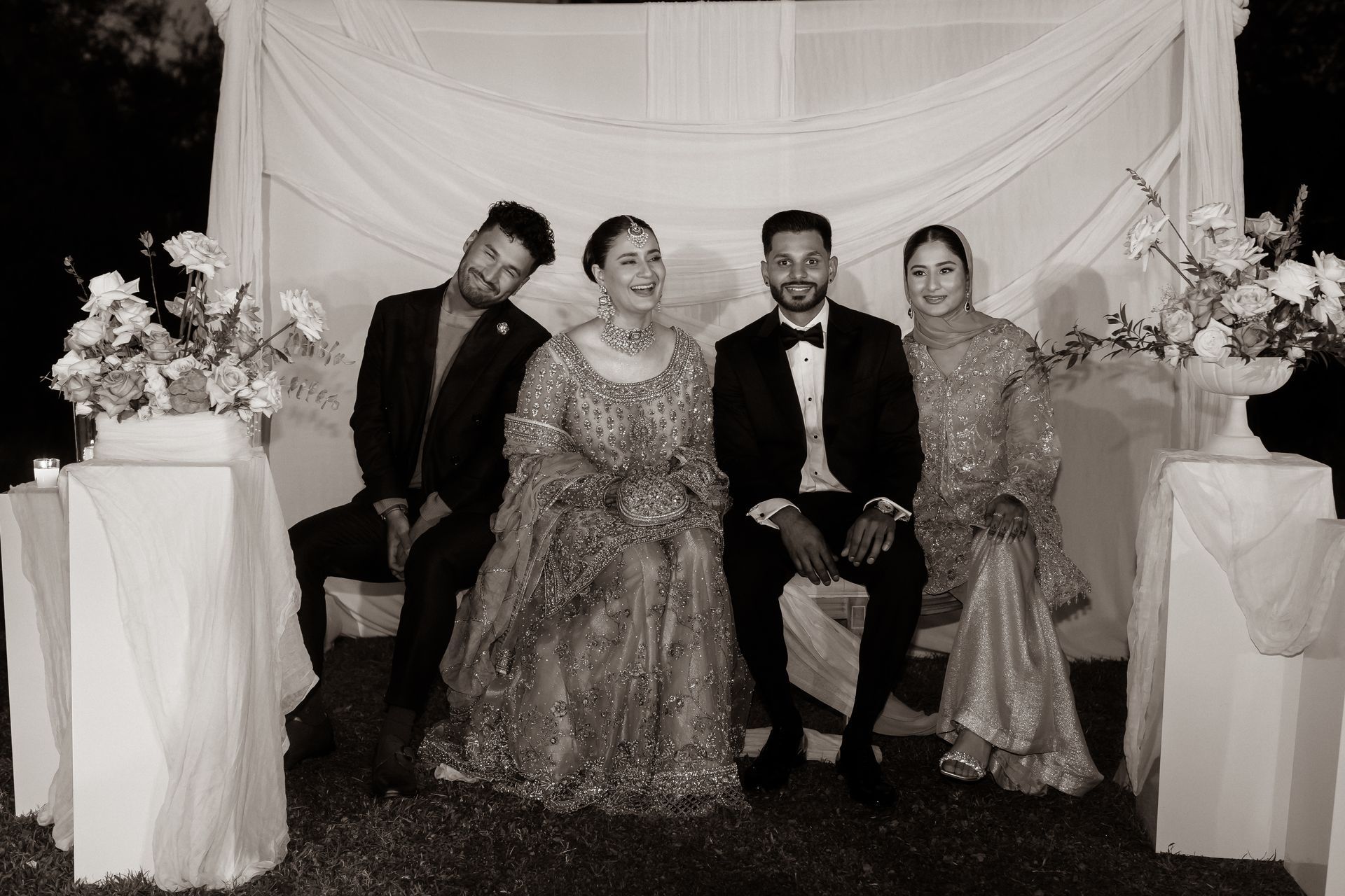 Four people pose for a formal photo. A woman in a gown sits with three others by a decorated backdrop.