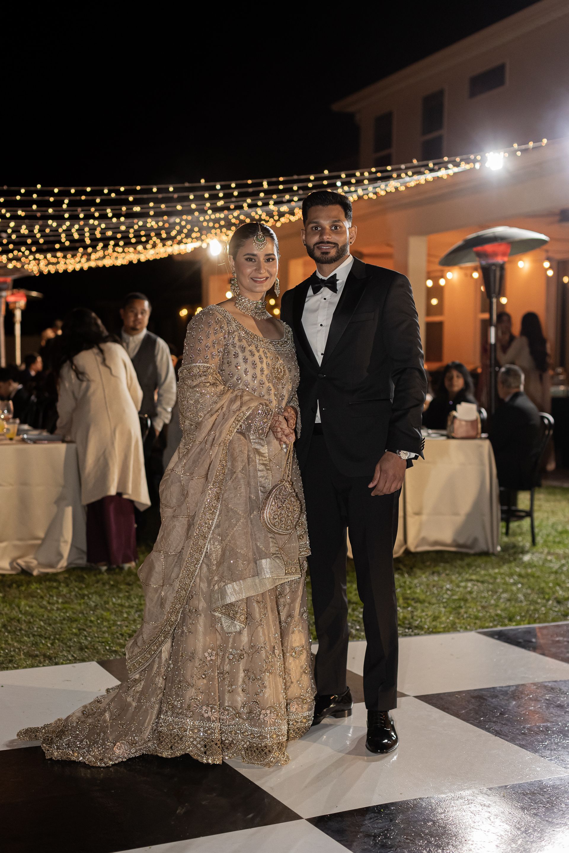 Couple in formal attire pose for a photo at an outdoor evening event. Gold sequin dress, black tuxedo.