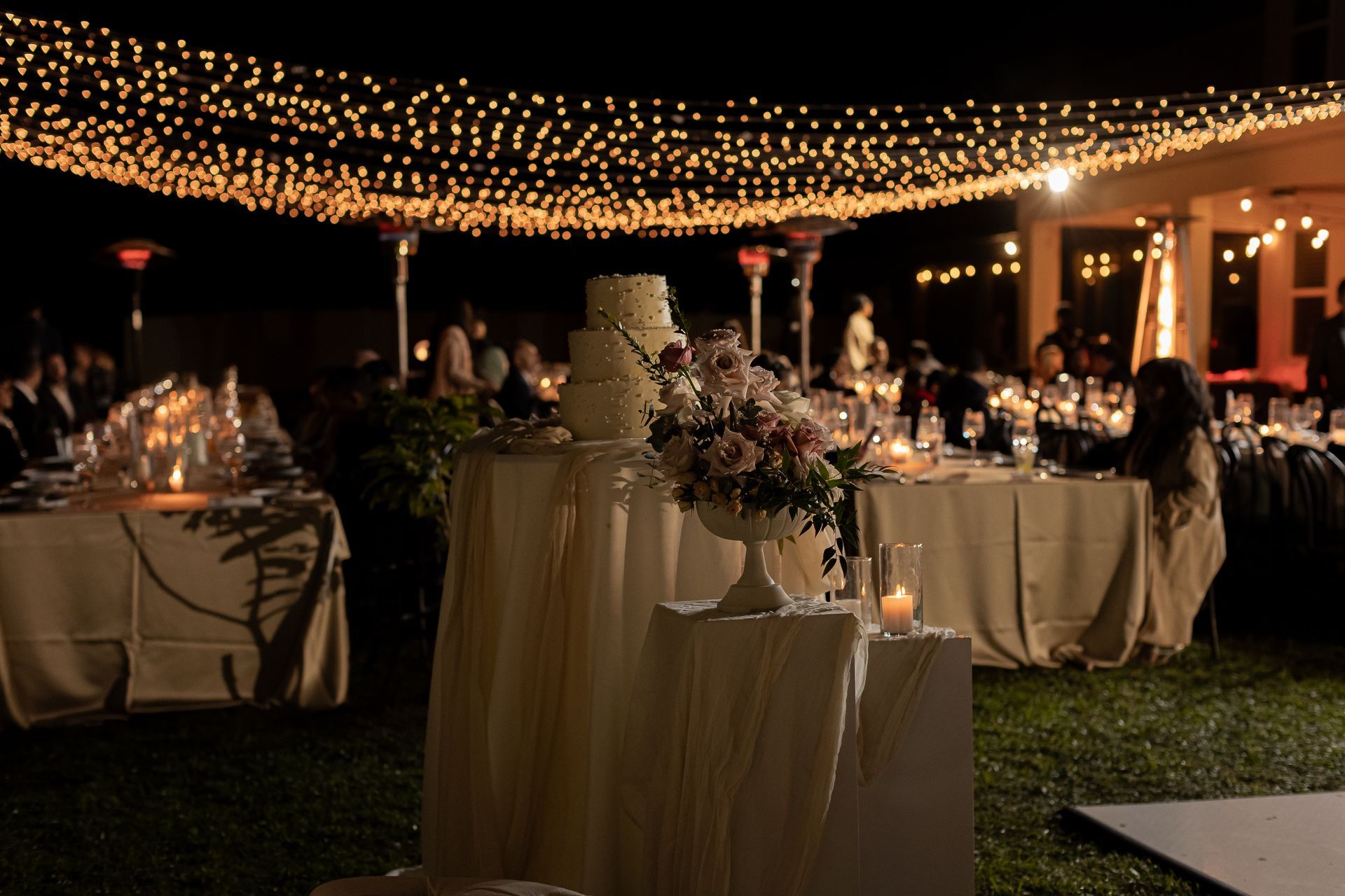 Wedding reception at night with cake, floral arrangement, and string lights.