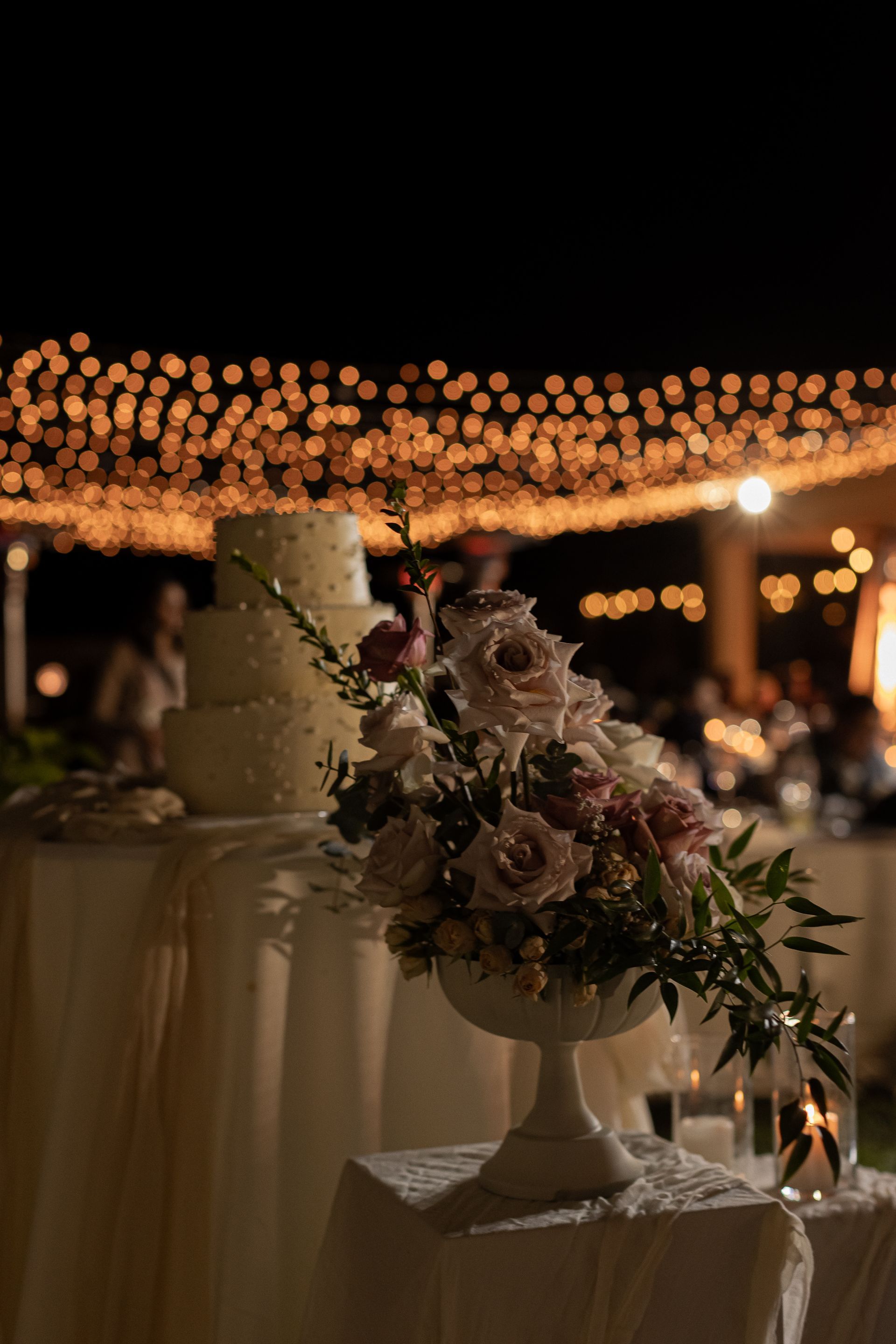Wedding reception at night with floral arrangement on a table, cake in the background, and string lights.