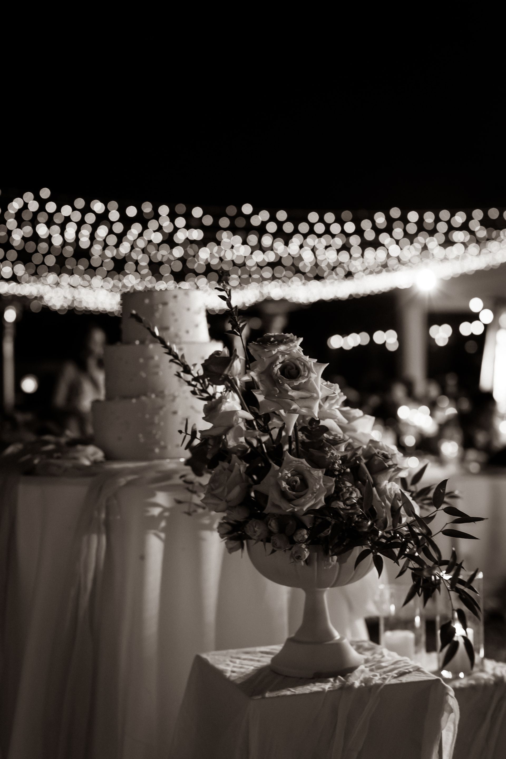 Wedding cake and floral arrangement under string lights at a night reception.