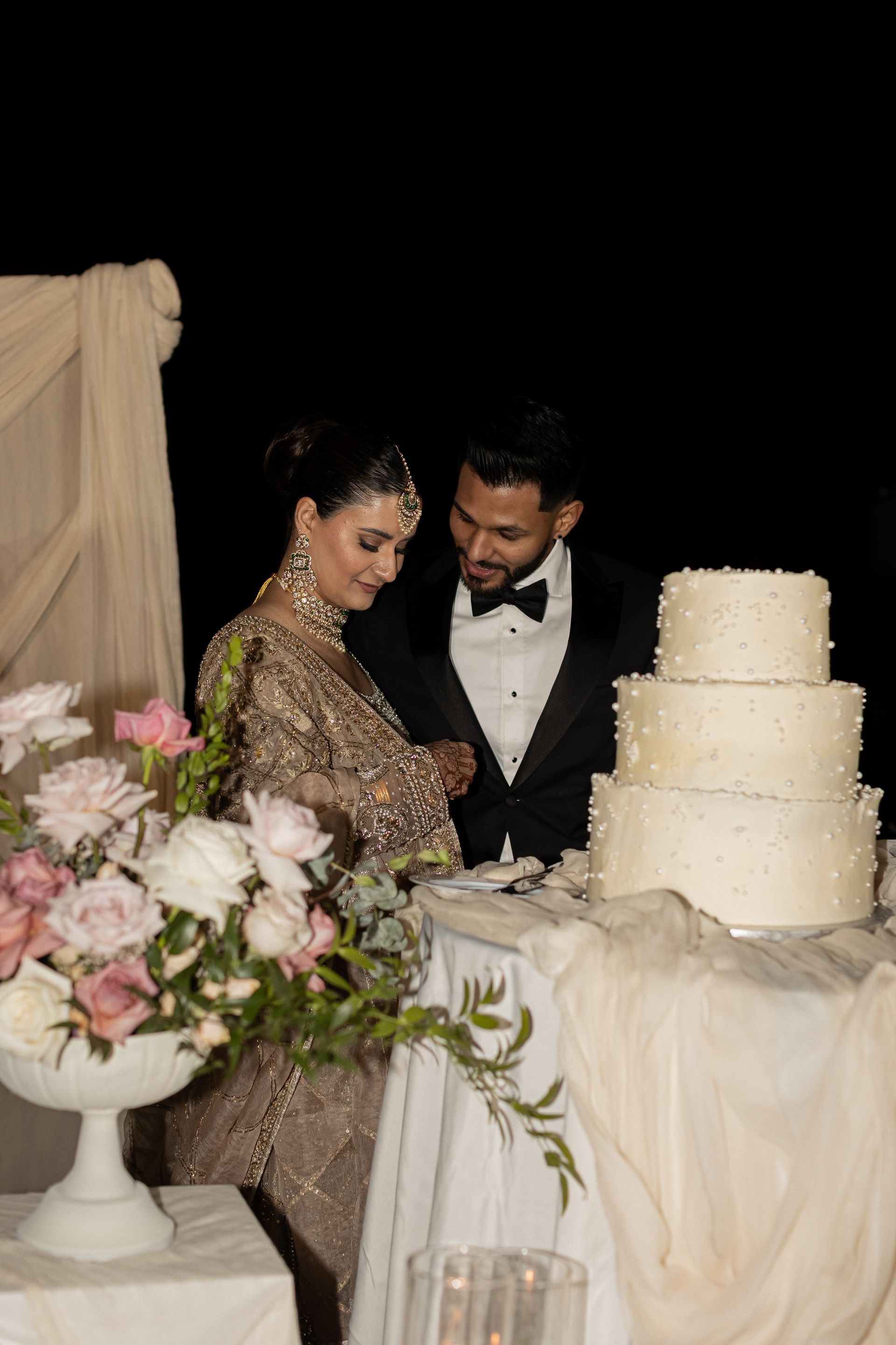 Couple cutting a tiered wedding cake at an outdoor reception. They are smiling, and florals are on the table.