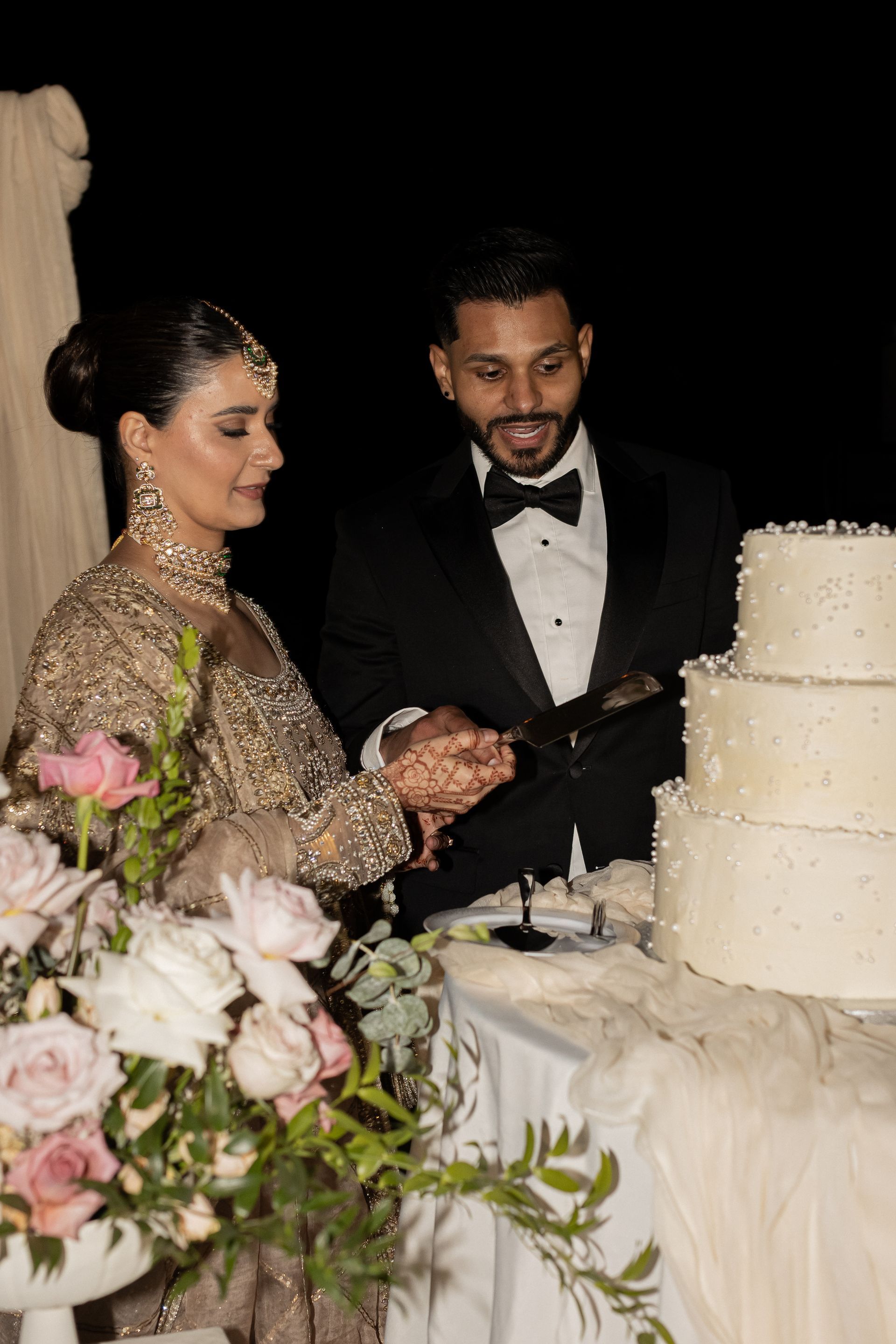Couple cutting a three-tiered wedding cake; woman wears embellished gown, man in tuxedo, flowers in foreground, at night.