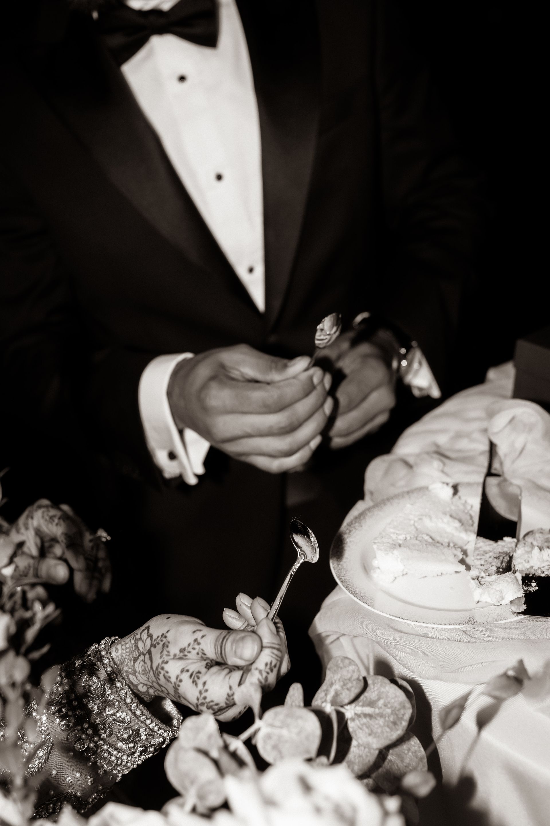 Groom in tuxedo cutting wedding cake with ornate utensils. Close-up, black and white.