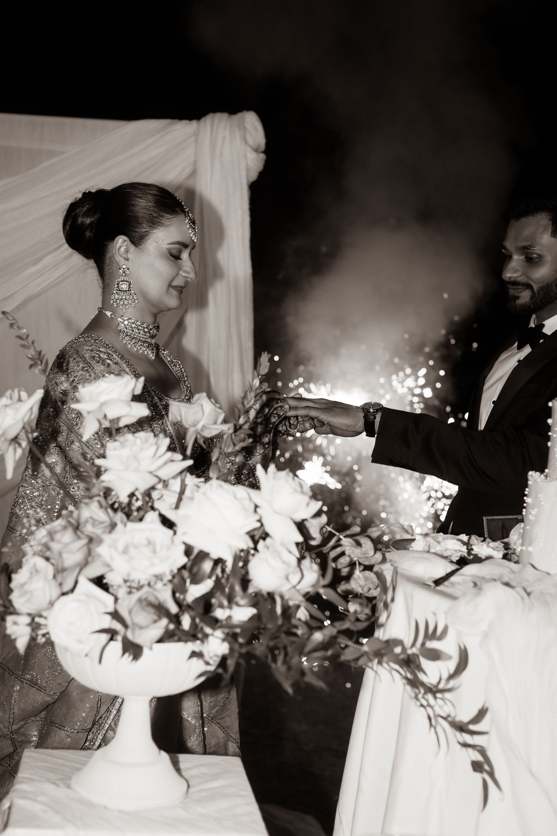 Bride and groom at a wedding, near a floral arrangement, with fireworks.