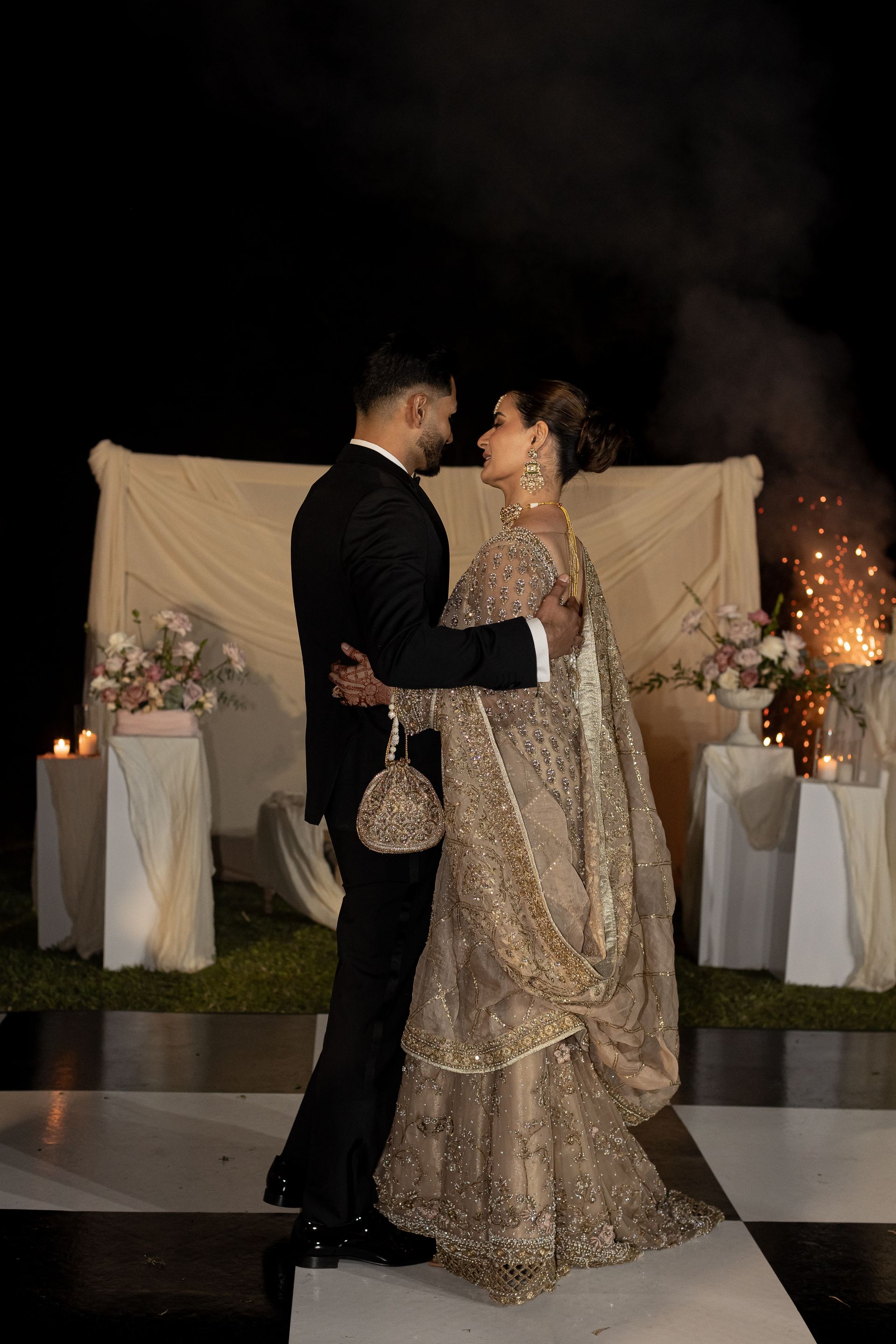 Couple dancing at an outdoor event, man in a tuxedo, woman in a gold gown, fireworks in the background.