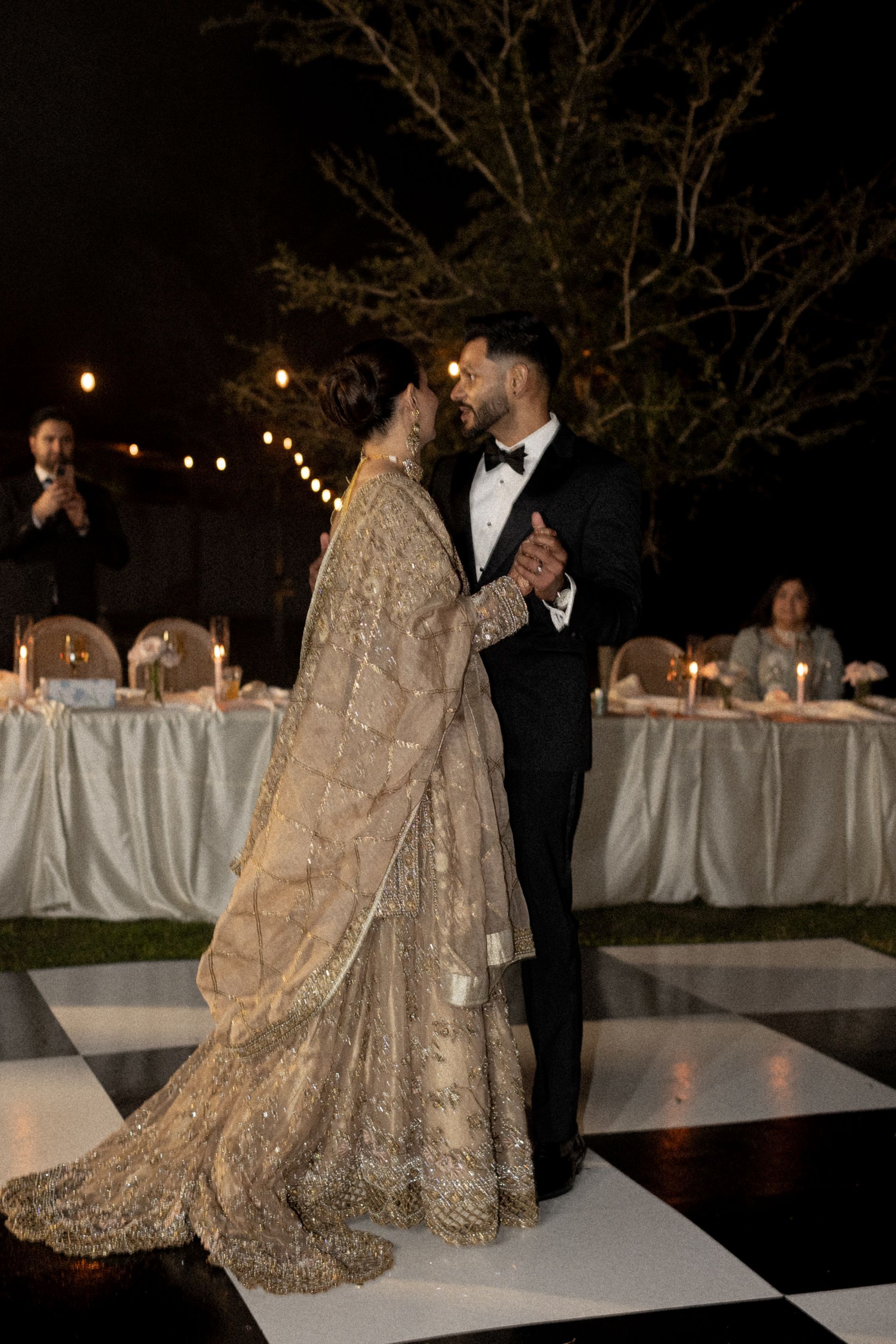 Couple dancing on checkered floor at night. Woman in gold gown, man in black tux, near decorated tables.