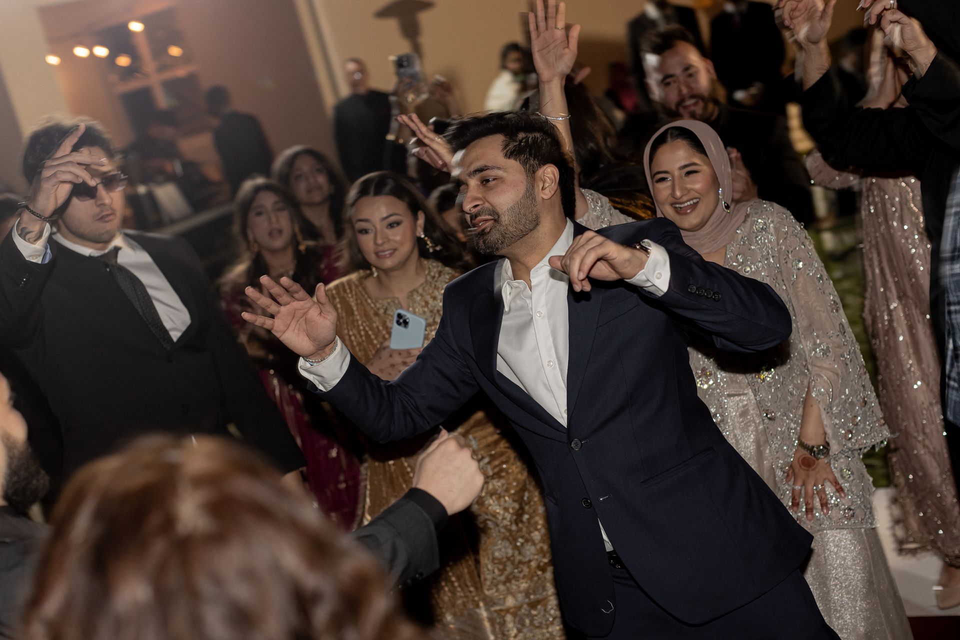People dancing at an event, man in suit center. Others in festive attire, arms raised, lively atmosphere.