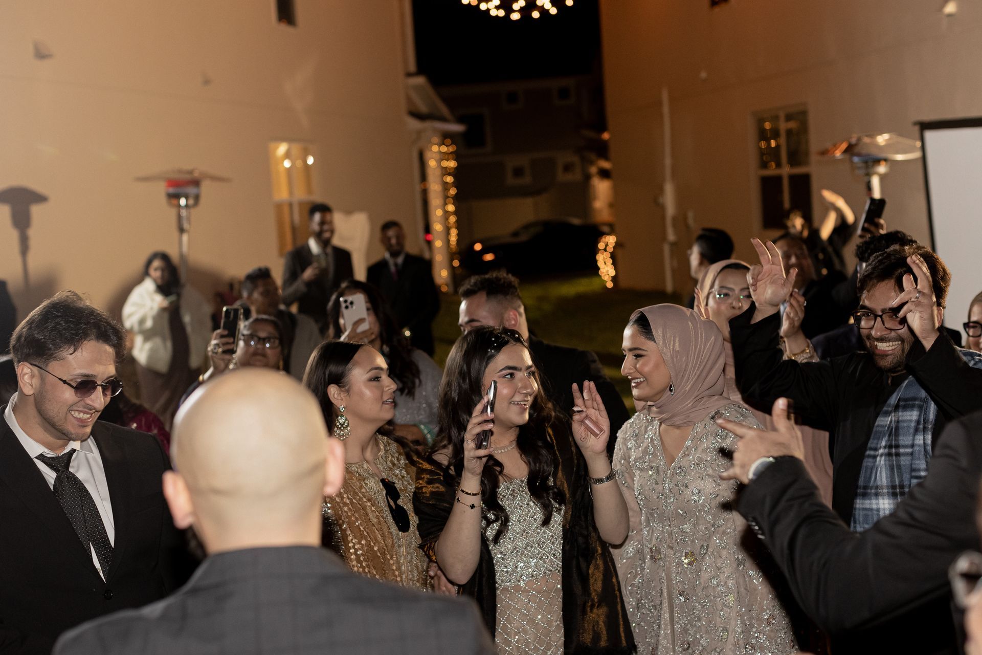 People dancing at an outdoor event with lights, wearing formal attire. Some are smiling, and one person is on the phone.