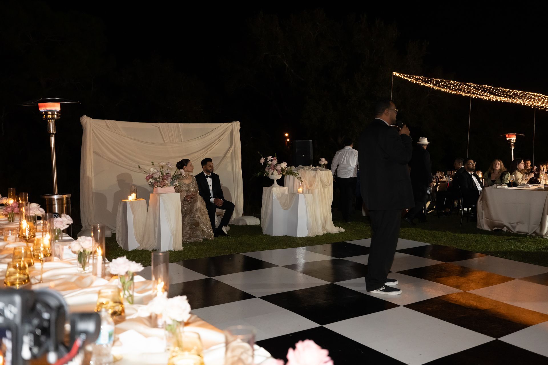 Nighttime outdoor wedding reception with a couple seated, a speaker, and guests at tables. Black and white dance floor.