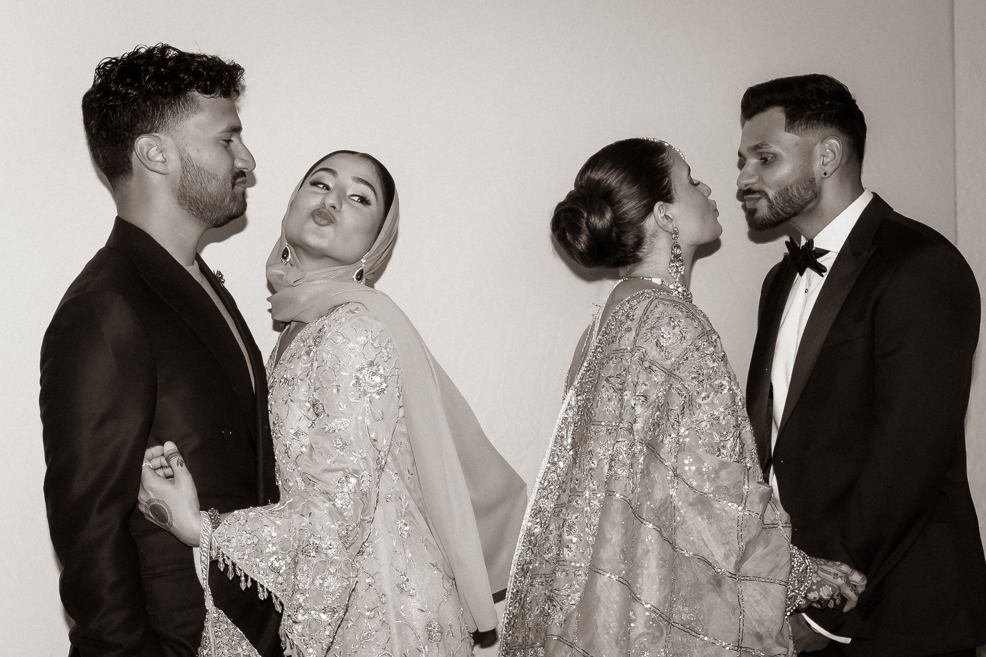 Two couples in formal attire pose cheek to cheek; one woman wears a hijab and embellished gown.