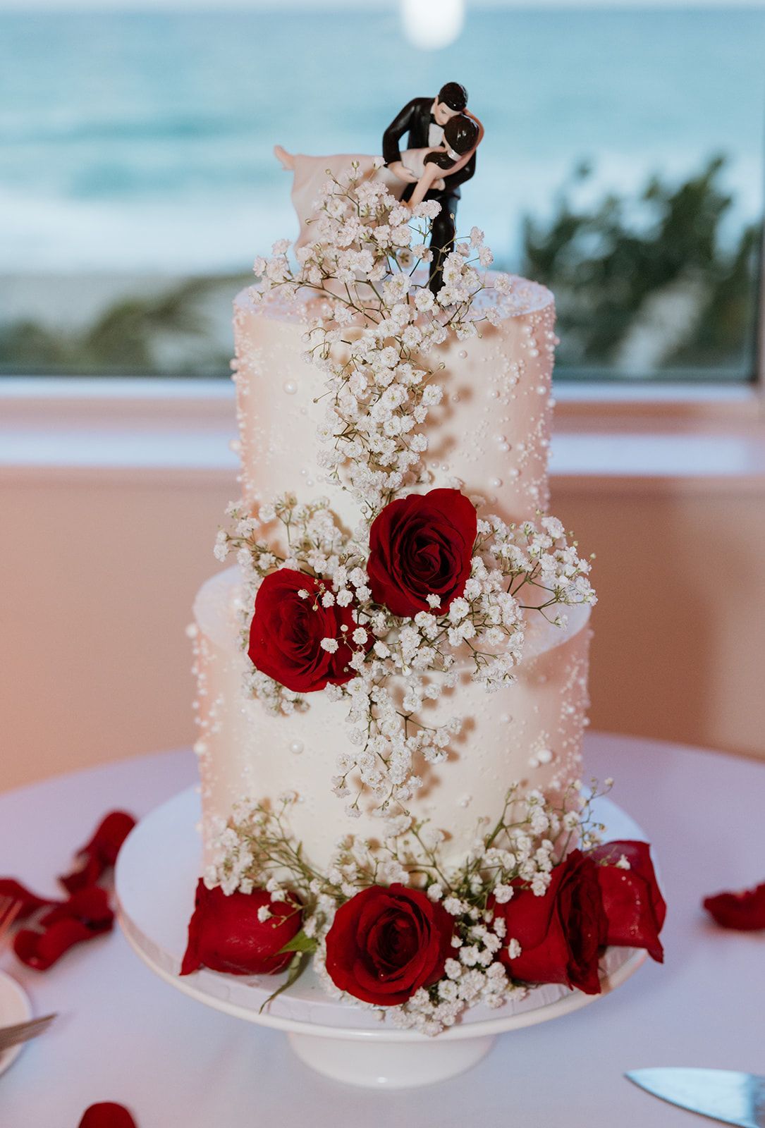 A wedding cake with red roses and baby 's breath on a table.