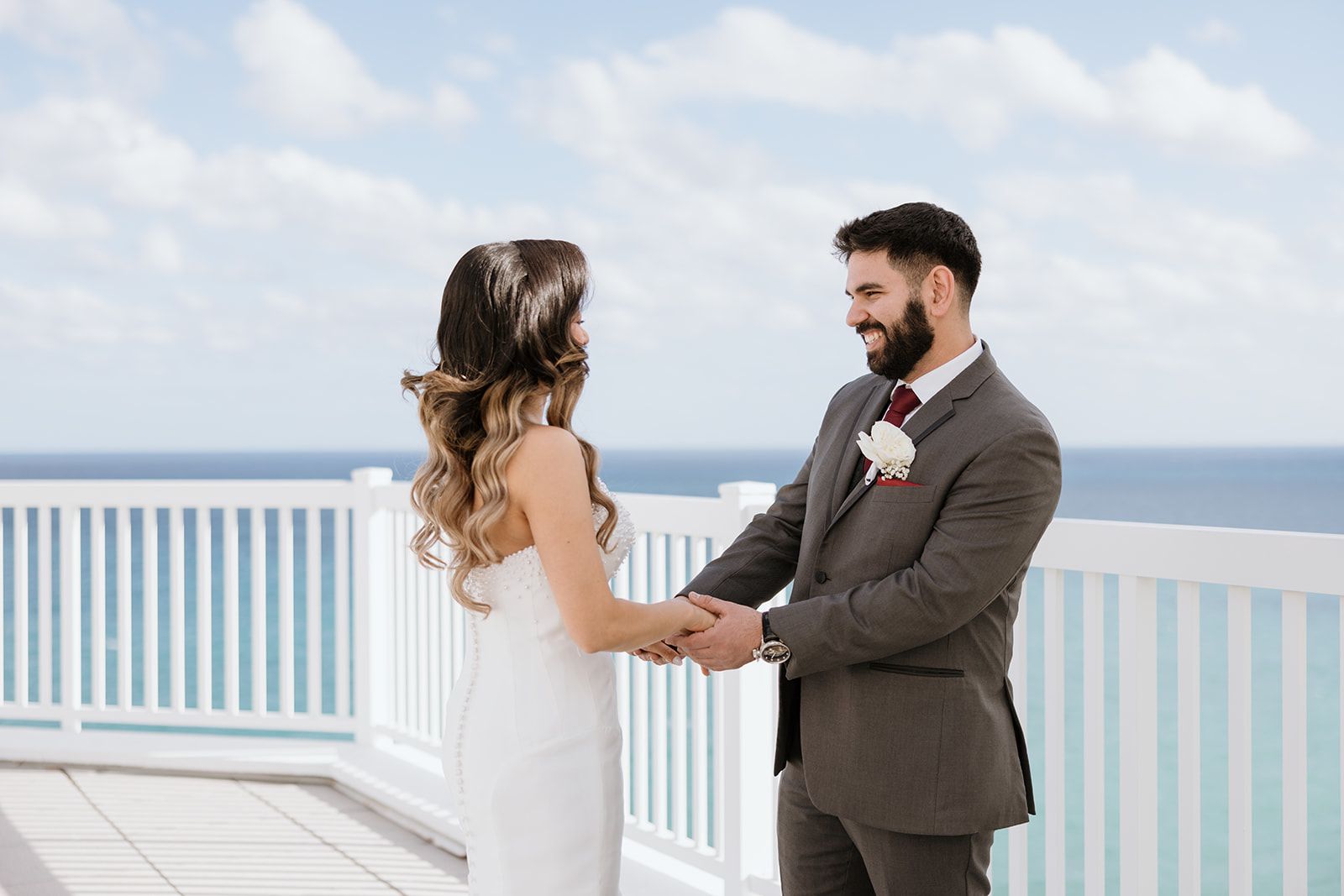 A bride and groom are holding hands on a balcony overlooking the ocean.