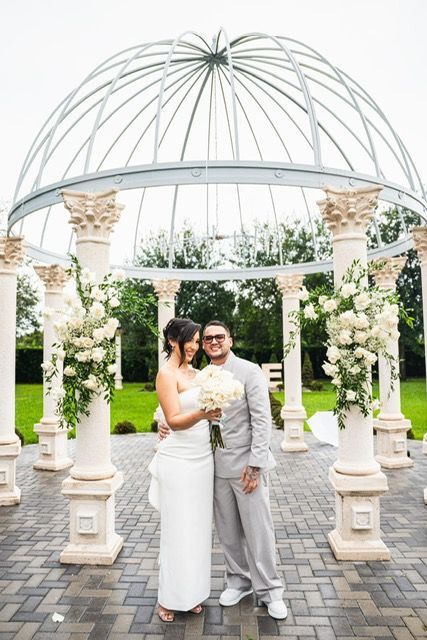 Bride and groom stand in front of a gazebo, she in white dress, he in gray suit, holding flowers.