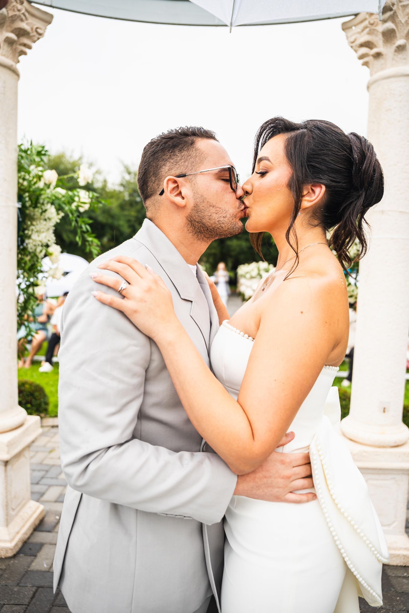 Couple kissing at outdoor wedding; bride in white gown, groom in gray suit.