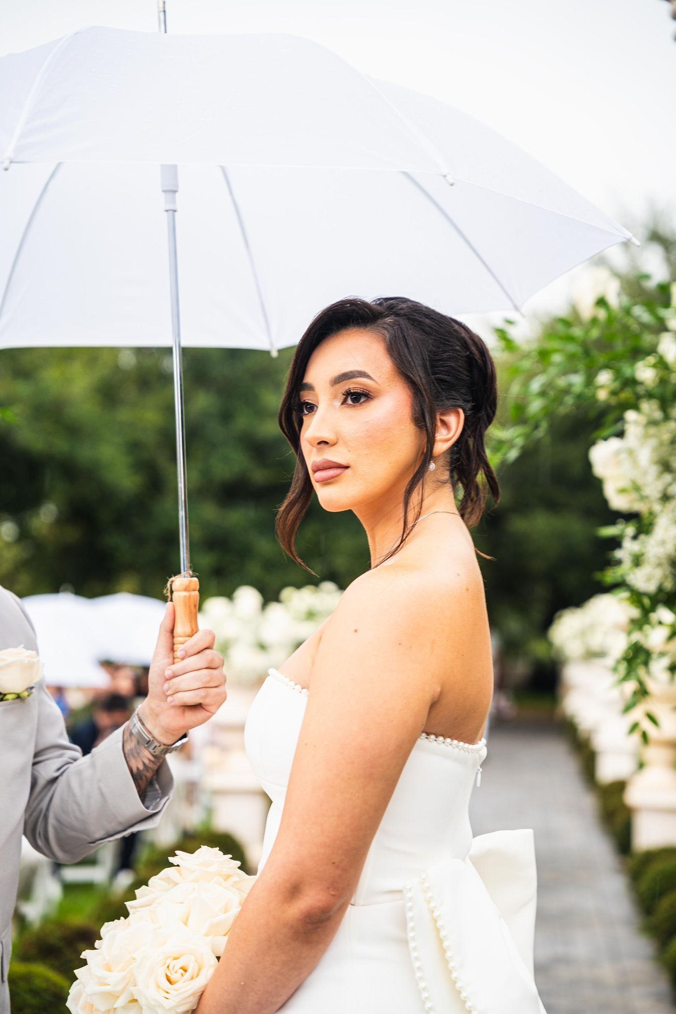 Bride under a white umbrella, looking away. Holding a bouquet, wearing a strapless gown; outdoor setting.