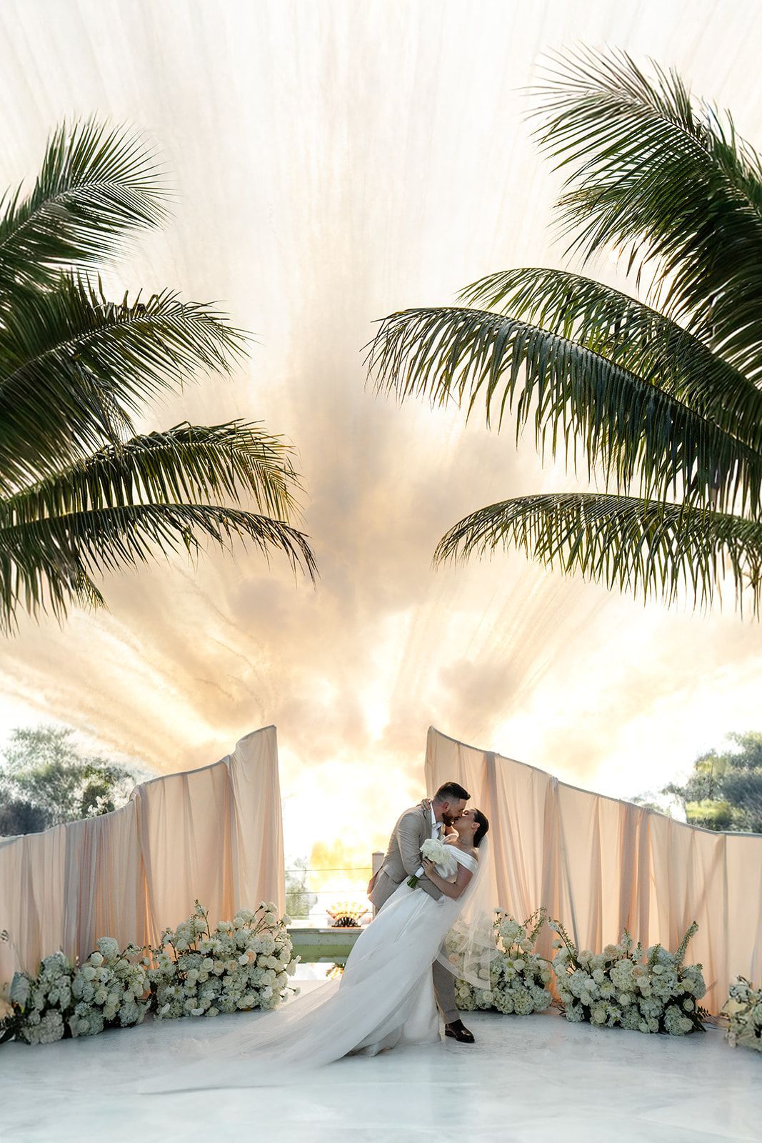bride and groom kissing while white white smoke fireworks are going off behind above the lake with ivory and cream wedding ceremony arch and flowers