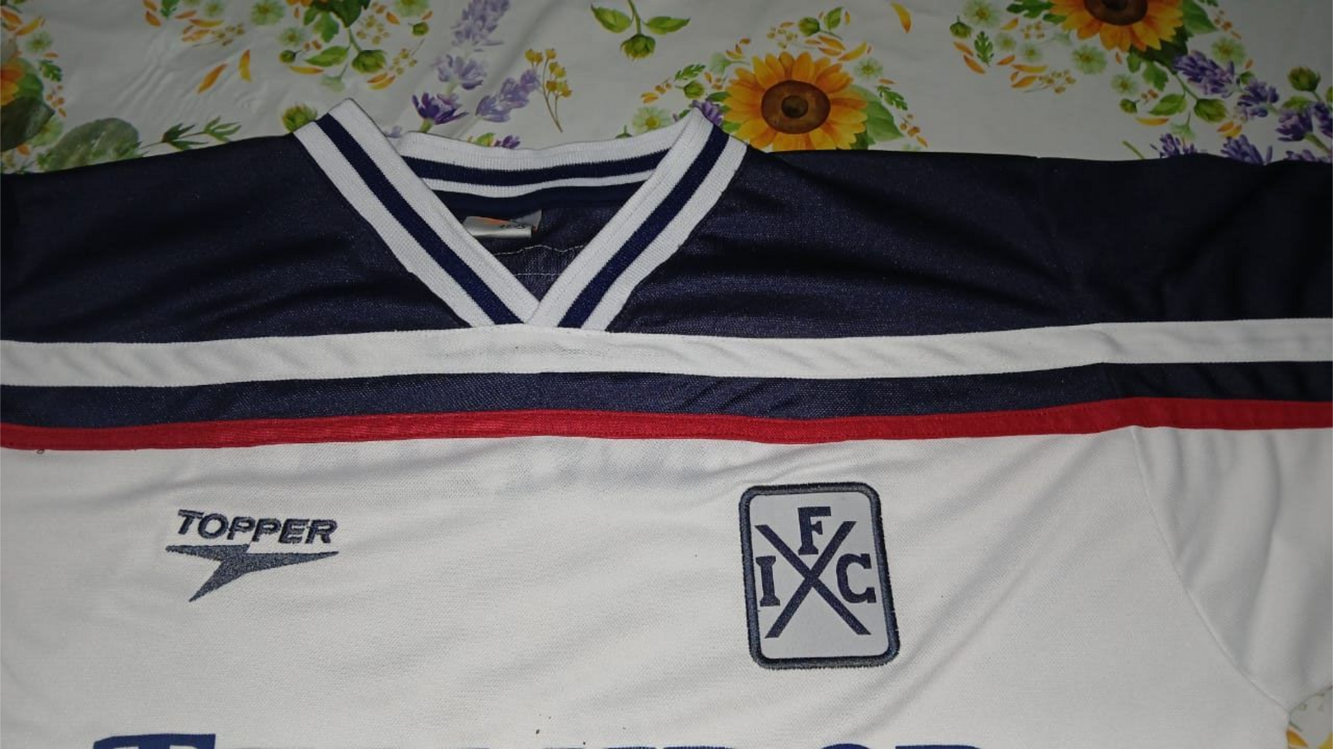 Camiseta de fútbol blanca con detalles en azul, rojo y blanco. Se ven el logotipo de