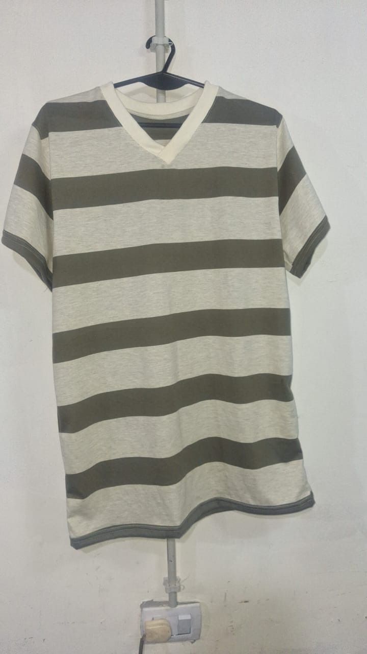 Camiseta con cuello en V, de rayas de color oliva y crema, colgada en un perchero frente a una pared blanca.