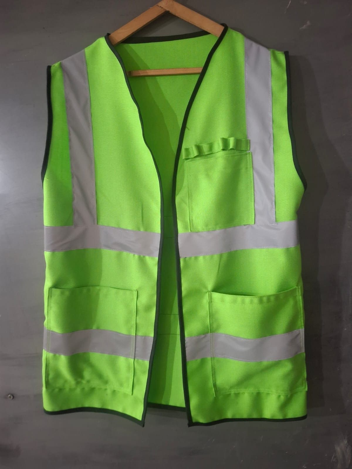 Chaleco de alta visibilidad de color verde con bandas reflectantes y bolsillos, colgado en una percha de madera.