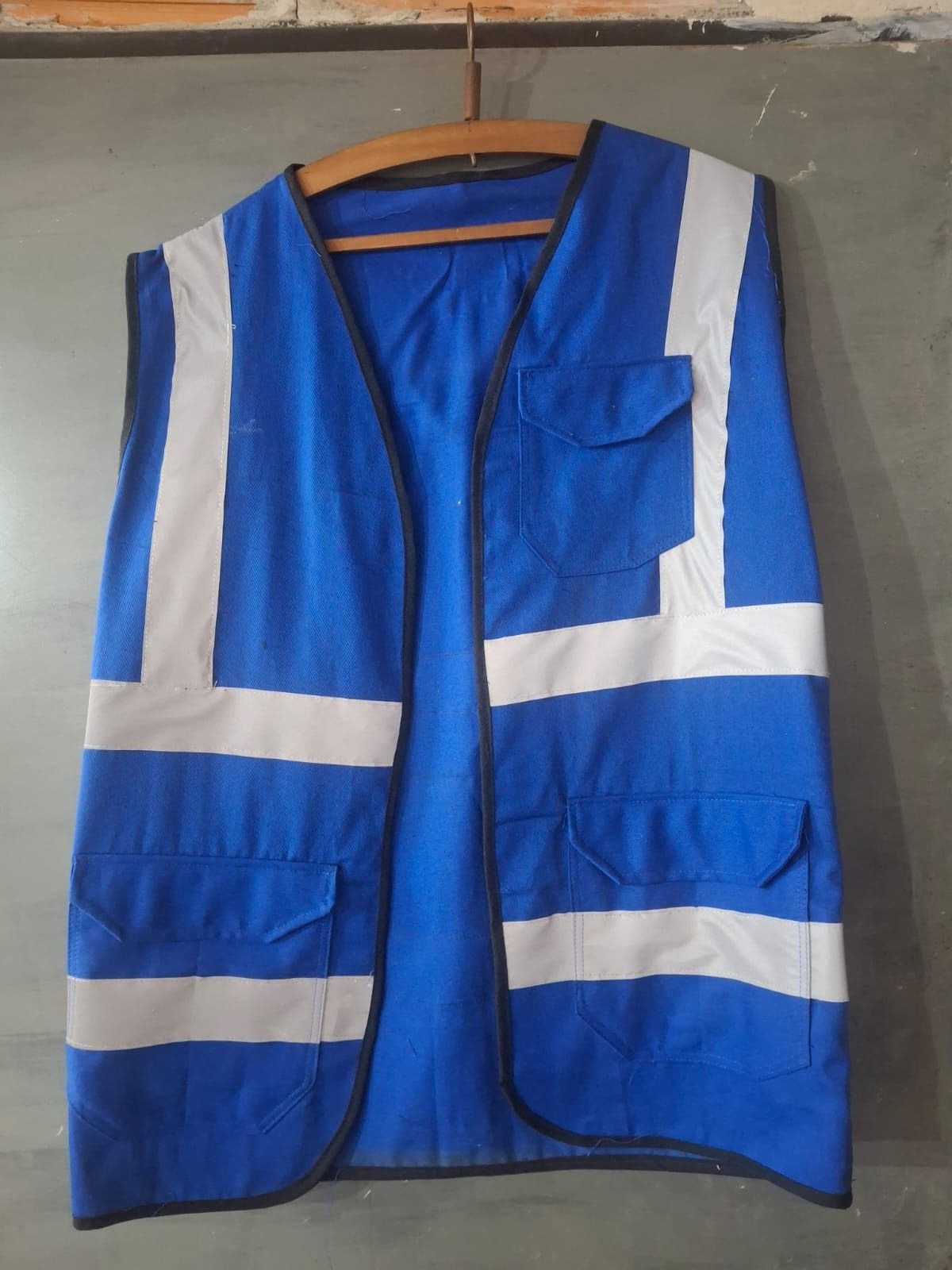 Chaleco de seguridad de color azul con franjas reflectantes blancas y bolsillos, colgado en una percha de madera.