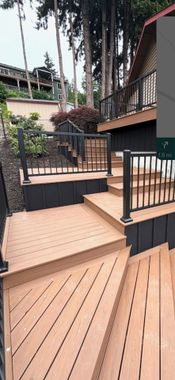 Wooden deck and steps with black railing leading to a house entrance, with a small tree and patio area nearby