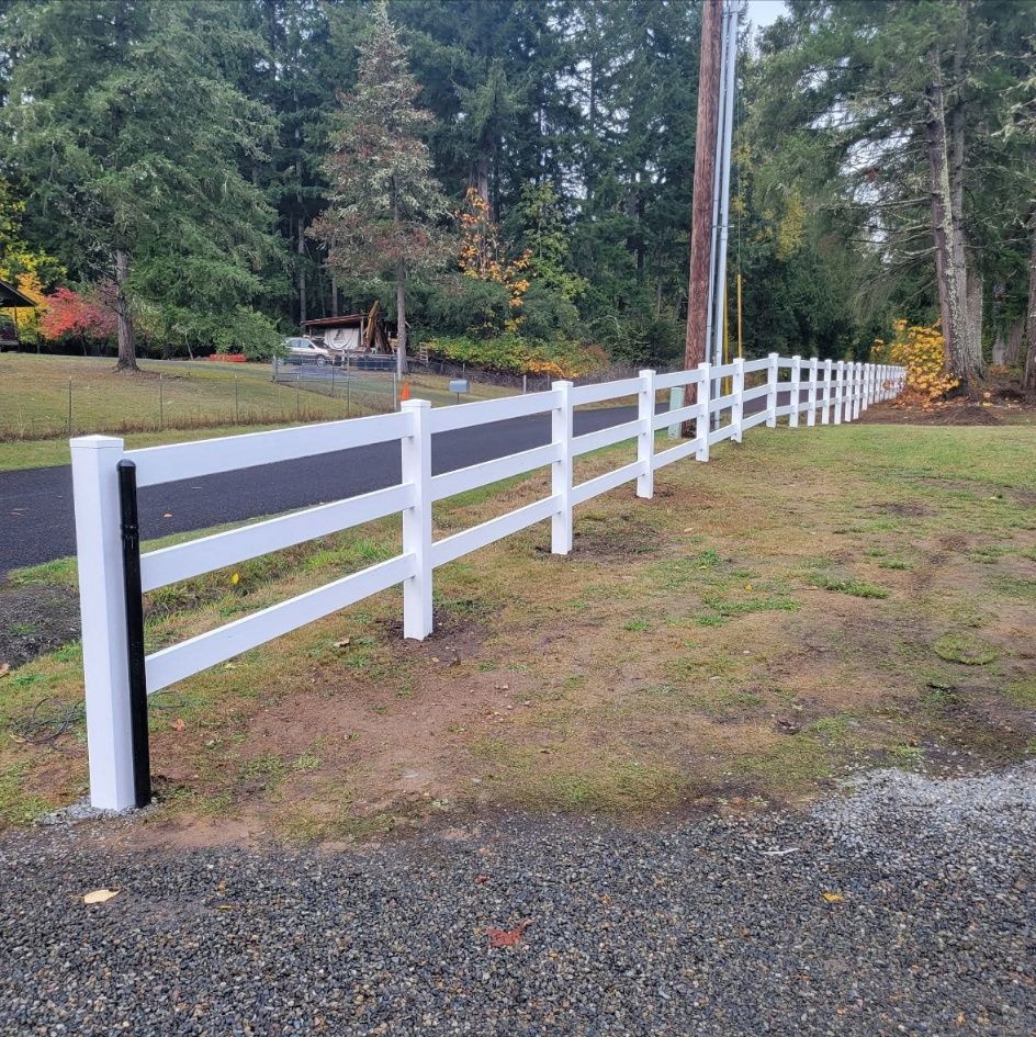 White wooden fence along a grassy roadside under tall trees, with gravel in the foreground.