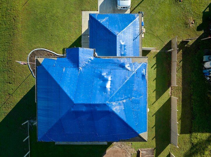 Aerial view of a house with a bright blue tarp covering their roof, surrounded by grass and a driveway.