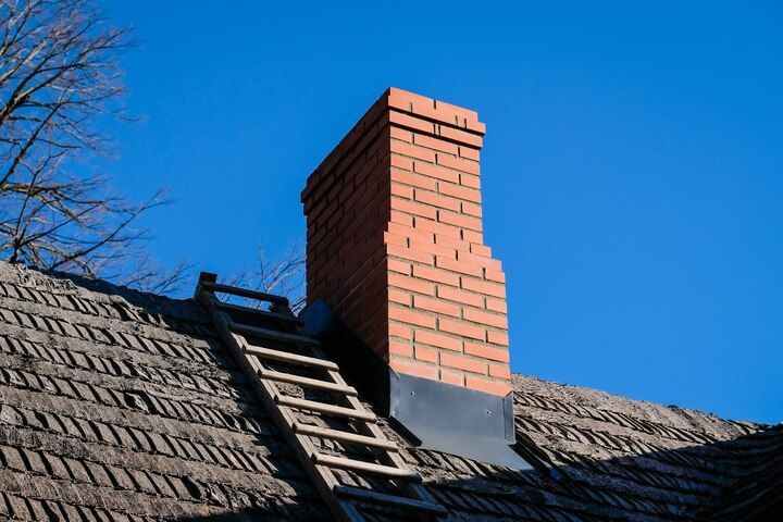 Brick chimney on a shingled roof with a ladder against it under a clear blue sky