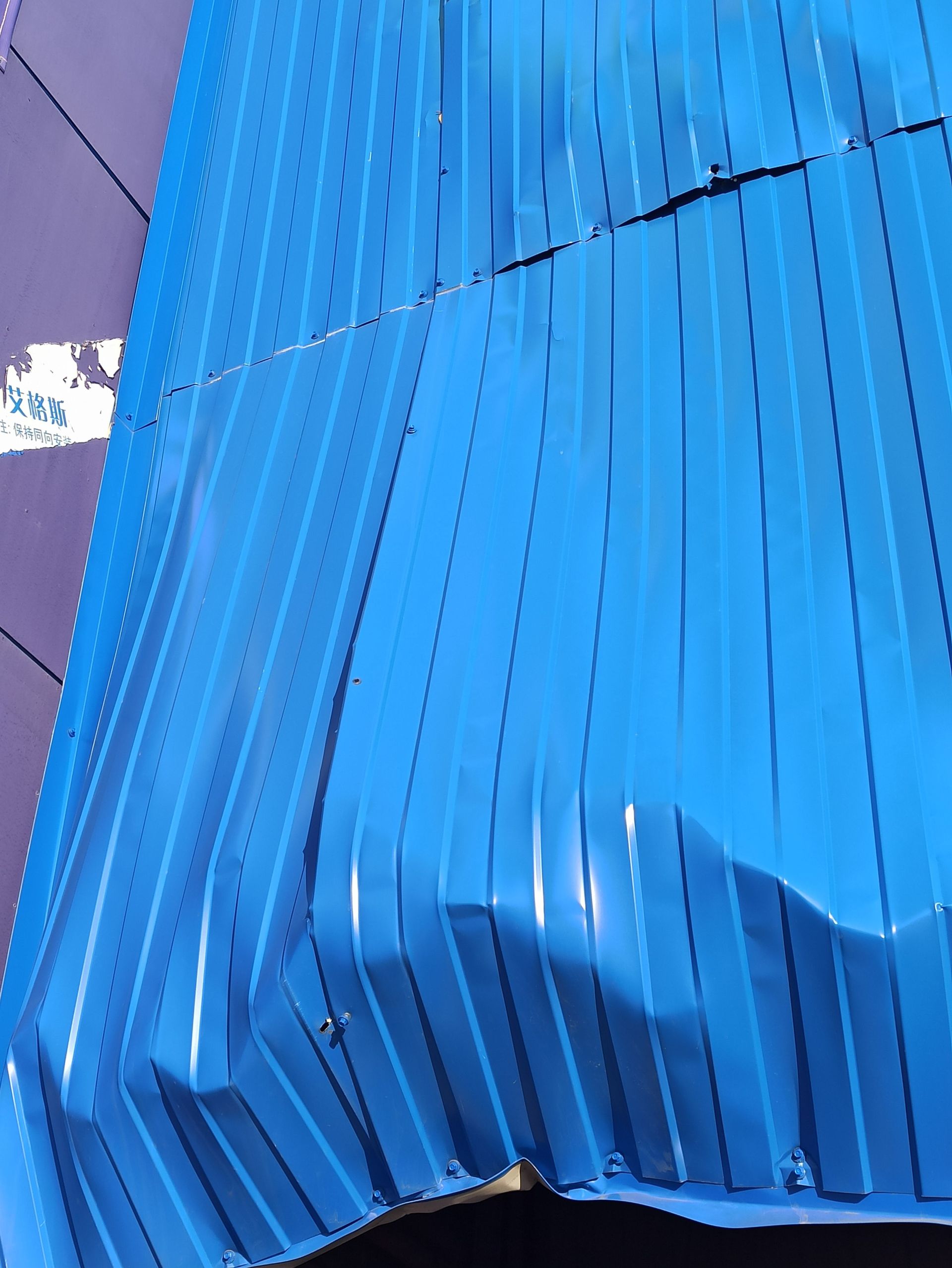 Blue corrugated metal wall or fence with a dented lower section against a purple background