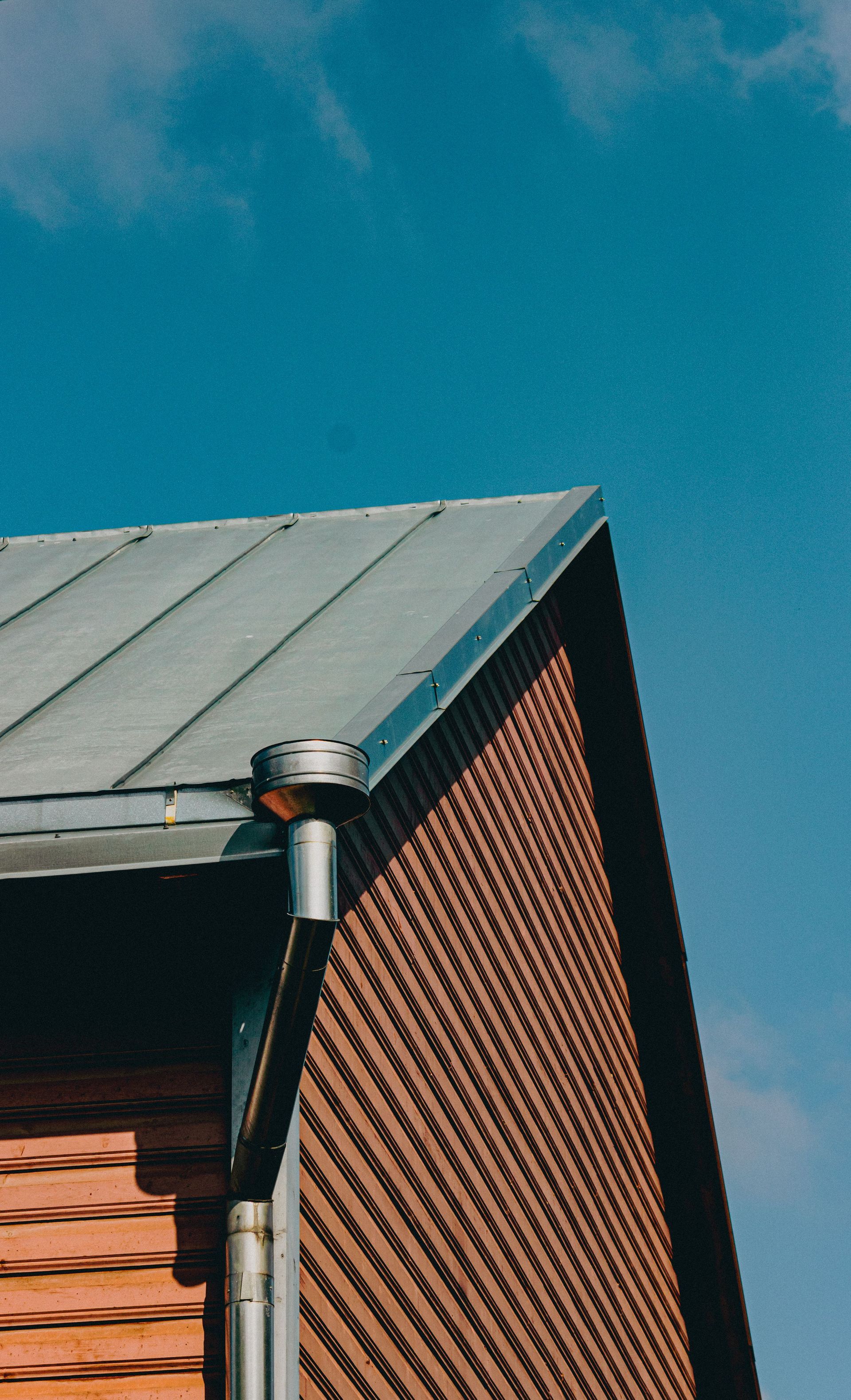 Brick house corner with gray metal roof and downspout against a blue sky