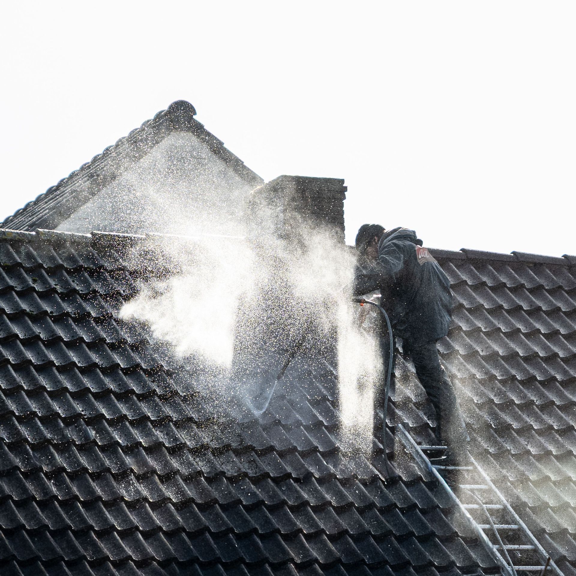 Workers power-washing a dark tiled roof, with water spraying over the shingles.