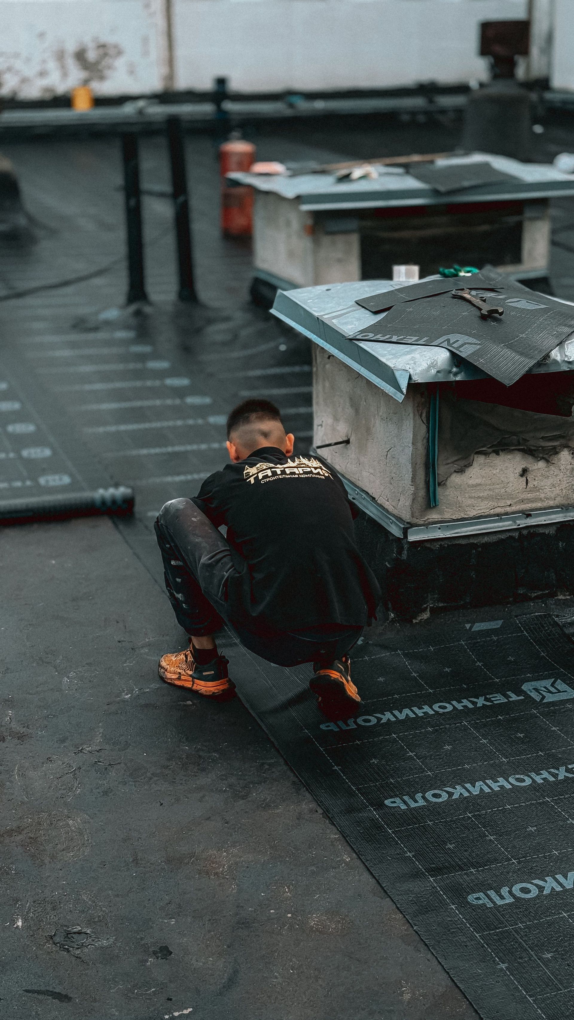 Person crouching on a wet rooftop beside a low concrete vent, wearing a dark jacket and orange shoes.