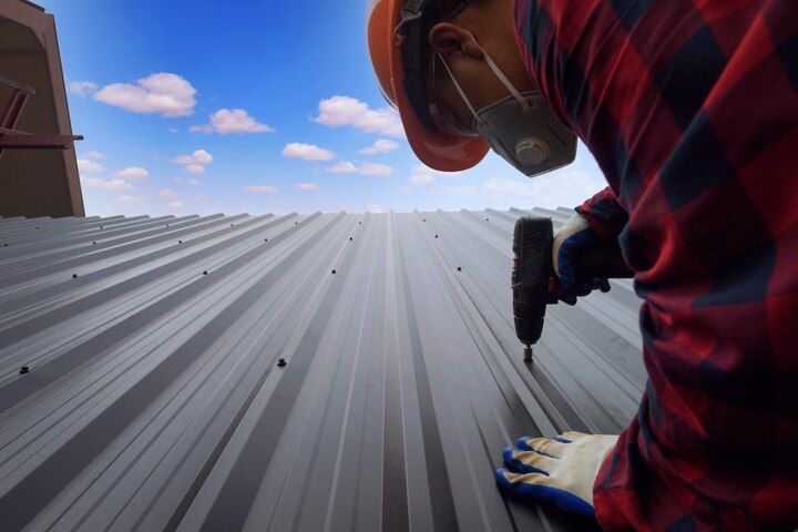 Worker in red plaid shirt installing a gray metal roof with a power drill under a blue sky