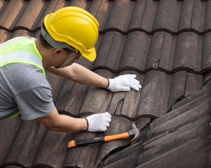 Worker in yellow hard hat repairing dark roof shingles with a hammer and gloves