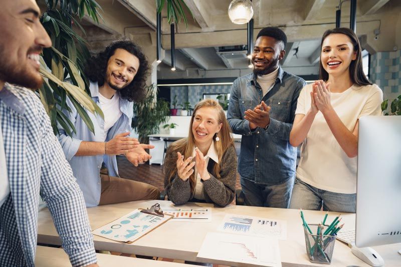 Group of people clapping at a desk in an office setting, smiling and looking at a person.