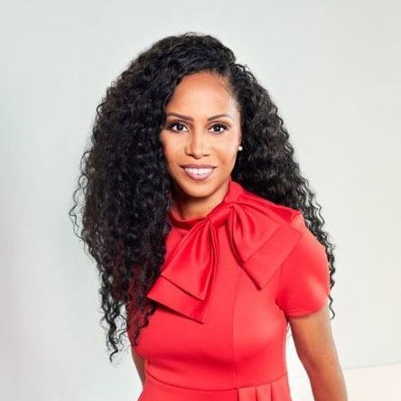 Woman with long, curly hair in a red dress with a bow, smiling, against a light background.
