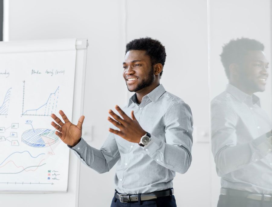 Man giving a presentation with animated gestures. Graphs and charts visible on a whiteboard in a bright room.