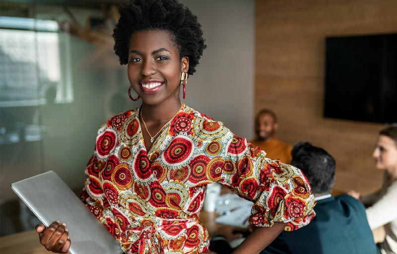 Woman smiling in office, holding laptop, wearing colorful print blouse, blurred colleagues in background.
