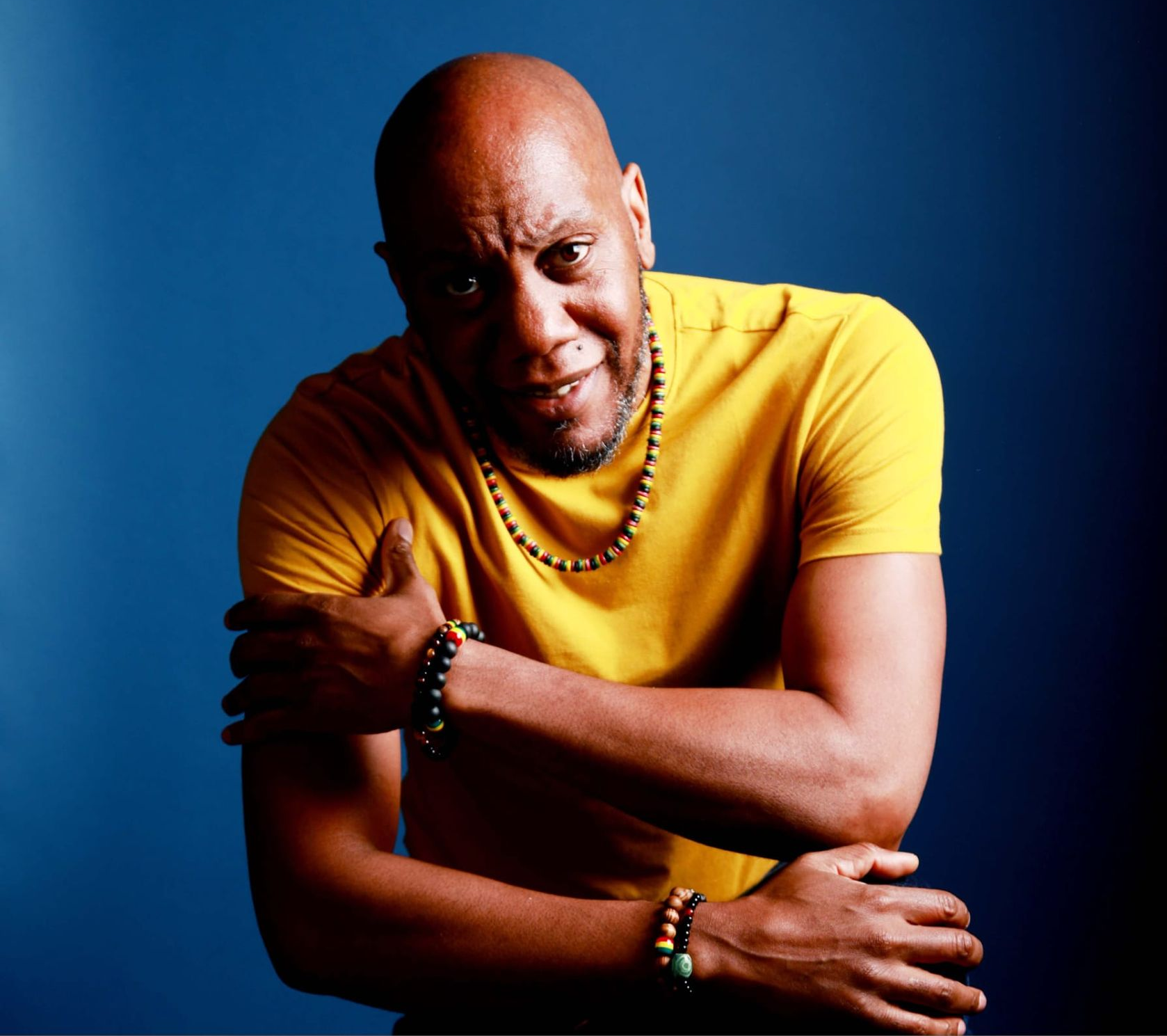 Man in yellow shirt, arms crossed, wearing beaded necklaces and bracelets, against a blue background.
