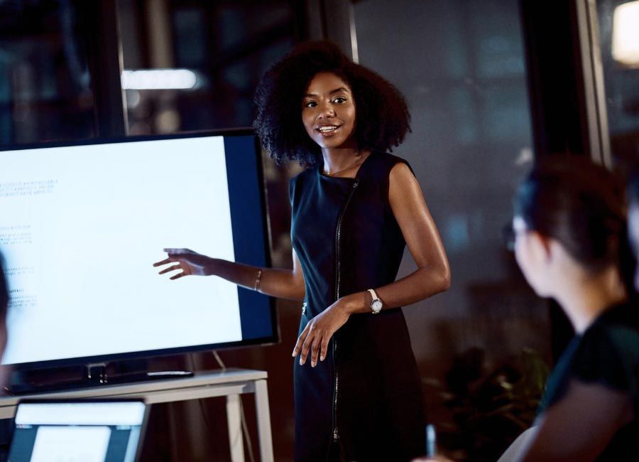 Woman giving a presentation with a smile, pointing at a screen in a modern office.