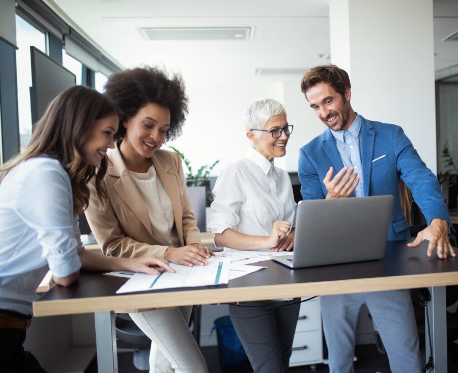 Four people collaborating around a laptop in a modern office, smiling, looking at the screen and documents.