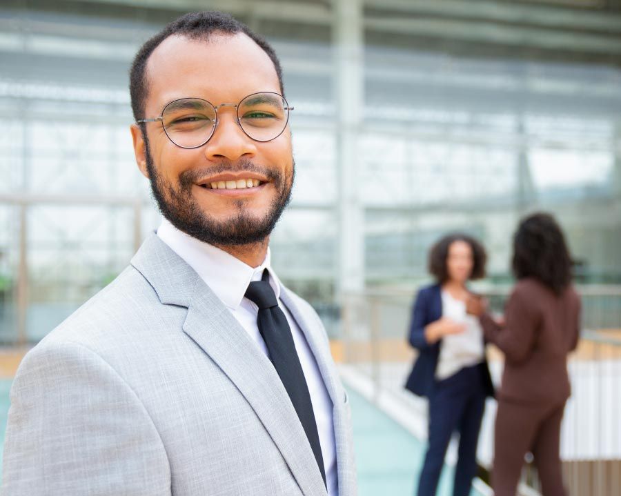 Man in suit smiling, with two women in suits blurred in the background. Outdoors, modern building.