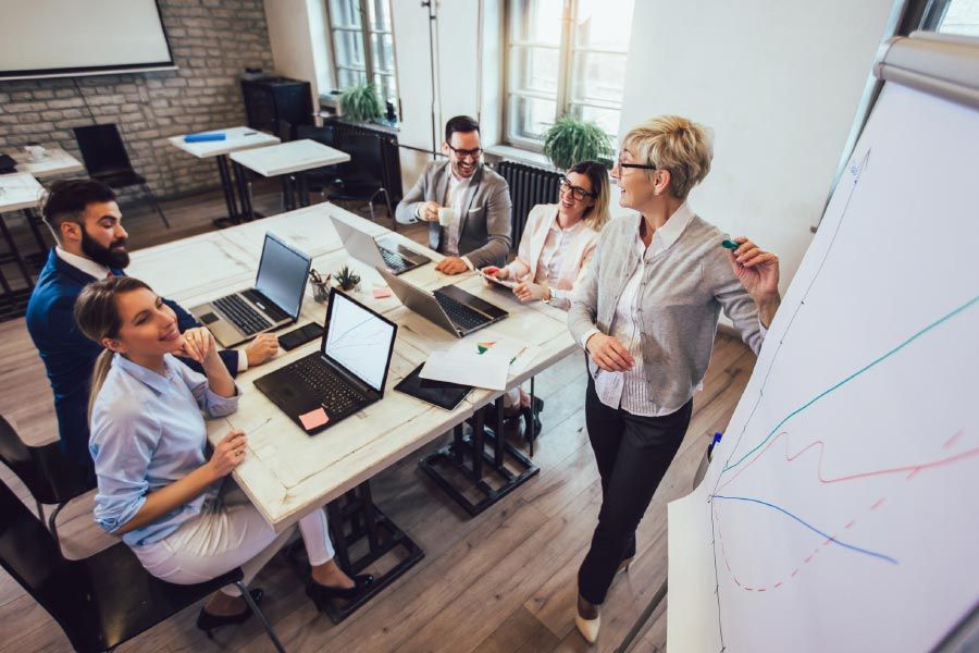 Business team in a meeting, woman presenting at whiteboard, others with laptops. Bright, modern office space.