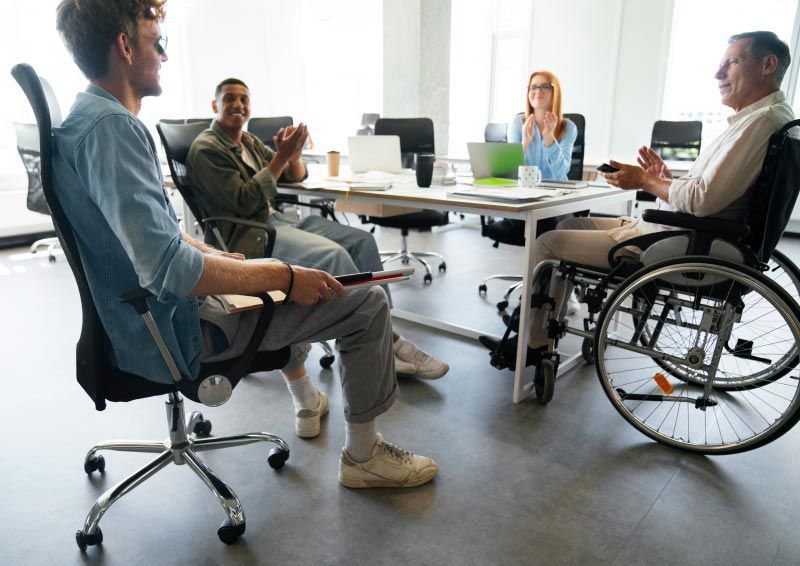 A diverse group in a meeting, with one person in a wheelchair. They are in a well-lit office, clapping and discussing.