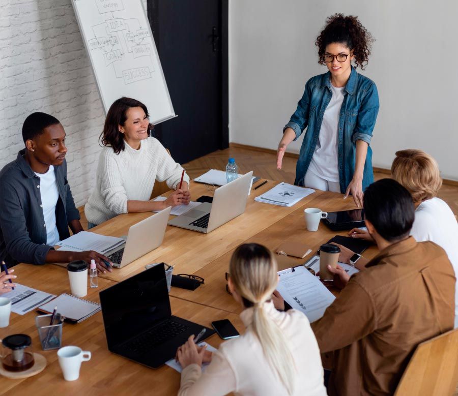 A team meeting at a wooden table; woman in denim jacket leads the discussion, others listen with laptops and papers.