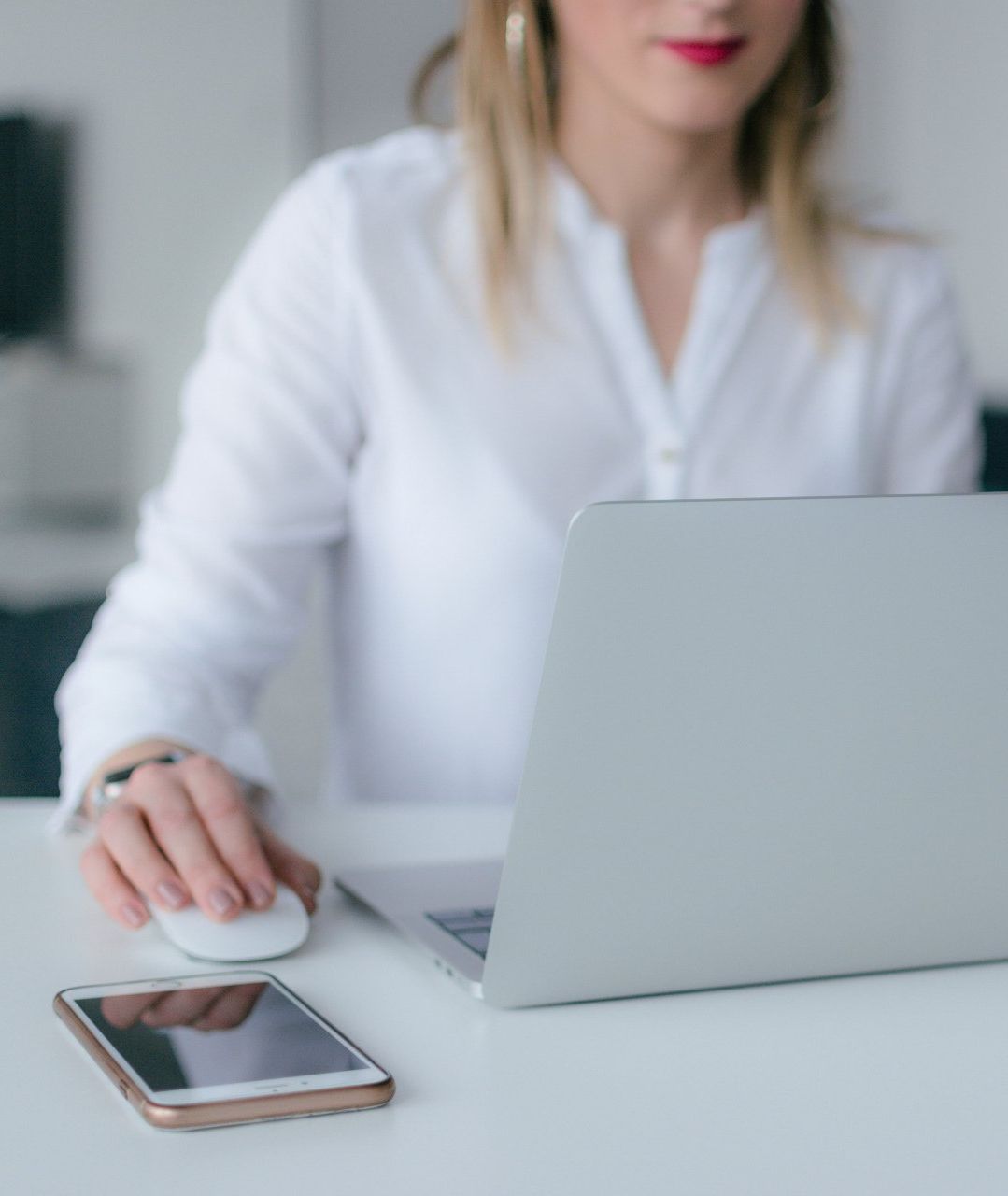 Woman in white shirt using a laptop and mouse at a desk, with a smartphone in the foreground.