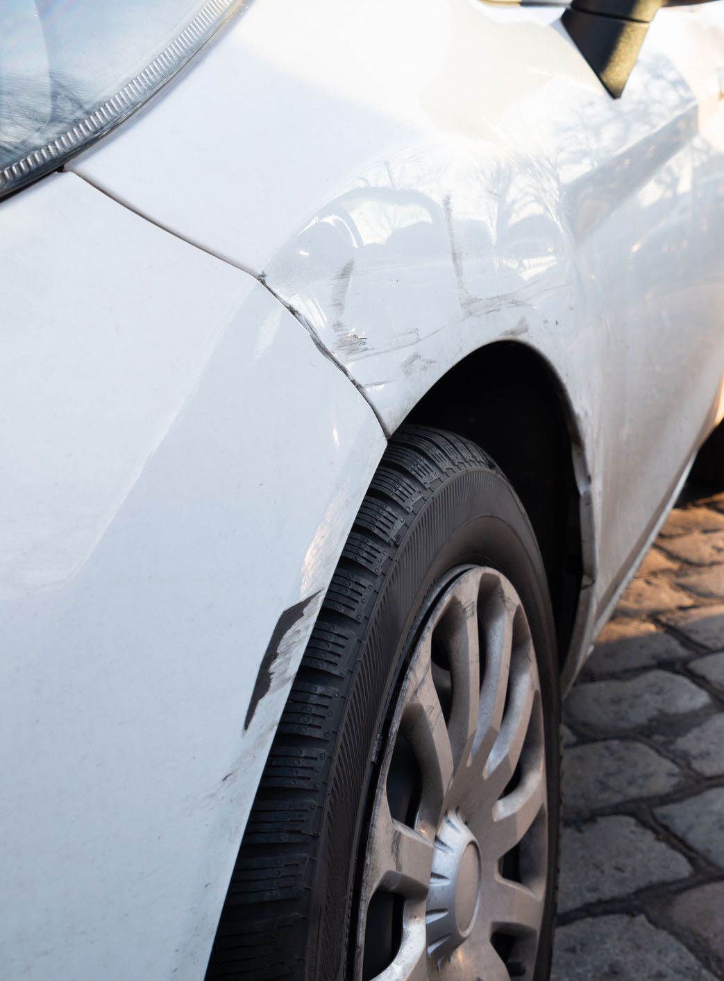 A white car with a damaged fender is parked on a cobblestone street.