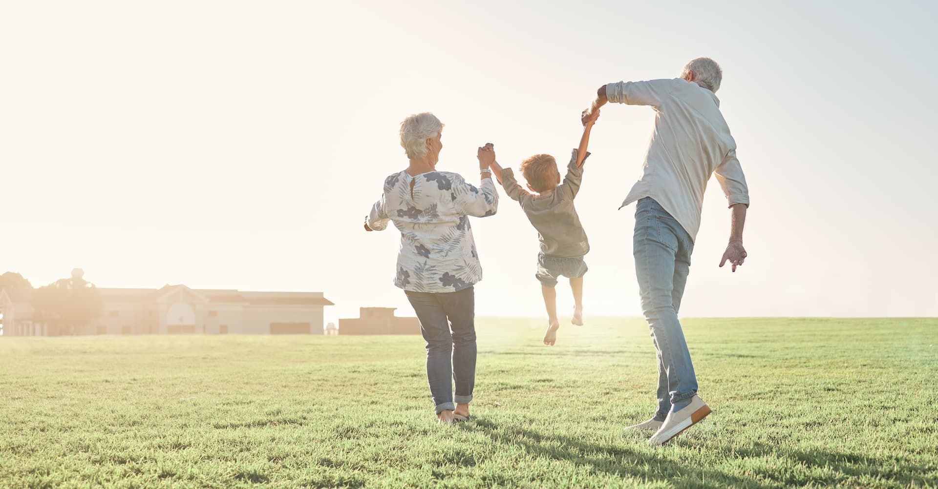 Two people hold hands with a child between them, lifting the child as they run through a grassy field in the sunlight.